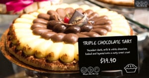 A triple chocolate tart sits on a plate. In front of it is a black tag with white writing displaying the name an price of the tart.