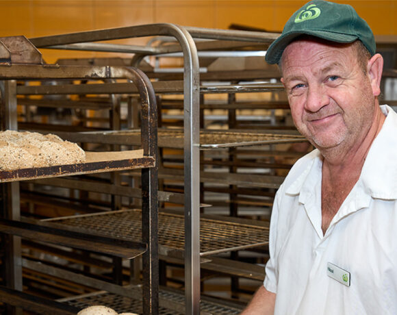 Woolworths Baker of the Year Nicol Strang is pictured in the kitchen. He's in a white shirt and green cap with the Woolworth logo on the front. He's holding a tray of buns, and there are metal shelves behind dhim.