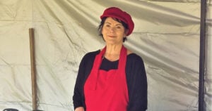 Cathy Hughes from Quince HQ stands a ta market stall. She wears a red apron and hat over a dark long sleeve shirt. Her dark hair is cut short.