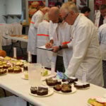 Judges at the BIEWA competition. Pictured are two men, one bald and one with grey hair and glasses. Both wear white coats. They are leaning over a table of baked goods.