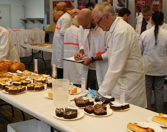 Judges at the BIEWA competition. Pictured are two men, one bald and one with grey hair and glasses. Both wear white coats. They are leaning over a table of baked goods.