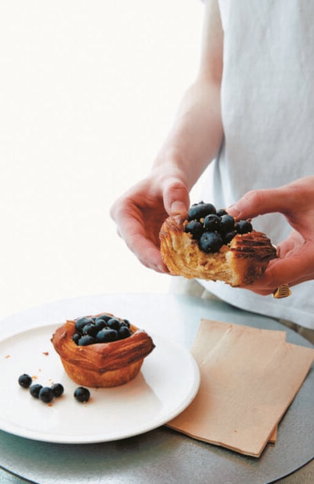 Baker Bleu's blueberry cheesecake danishes. Pictured is a complete danish on a white plate. There are a few loose blueberries on the plate around it. A person in a white shirt stands to the side holding a partially eaten danish.