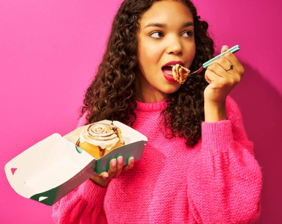 Cinnabon Australia has announced its expansion into WA. Pictured is a woman with long dark, curly hair. She wears a vibrant pink jumper and stand in front of a bright pink wall. She'sholding a blue Cinnabon branded box, with a scroll inside. She holds a fork with a bite of the pastry on it.