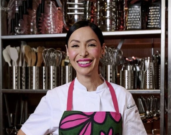 Katherine Sabbath has been named as a MasterChef Dessert Masters contestant. She's pictured standing in front of a wall of utensils. She wears a white long sleeved shirt and pink and green apron over the top. Her dark hair is tied back and she wears bright pink lipstick