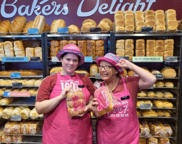 Kyabram Bakers Delight have been recognised as the top charity earners for the Pink Bun Campaign. Pictured are two female employees standing in front of a large wall of bread shelves. They each wear a pink top hat, pink apron and maroon Bakers Delight t-shirt. They're smiling at the camera, and each holding a bag of the pink buns.