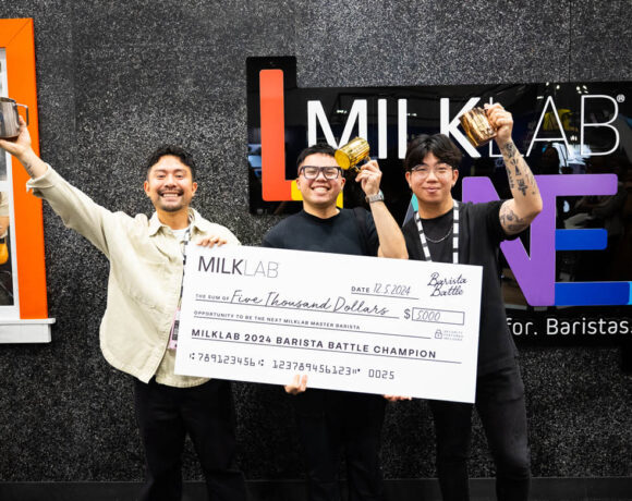 MILKLAB Barista battle winners. The trio are pictured in front of a grey wall, with the MILKLAB sign hanging on it. They hold a giant cheque in front of them and hold milk jugs over their heads.