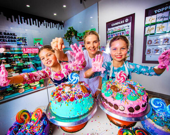 The Cheesecake Shop and Make-A-Wish Australia cake decorating workshop. Pictured are two young girls with blonde hair tied back. They've got icing on their hands, which they reach towards the camera, anbd two brightly decorated cakes in front of them. Their mum stands between them, smiling.