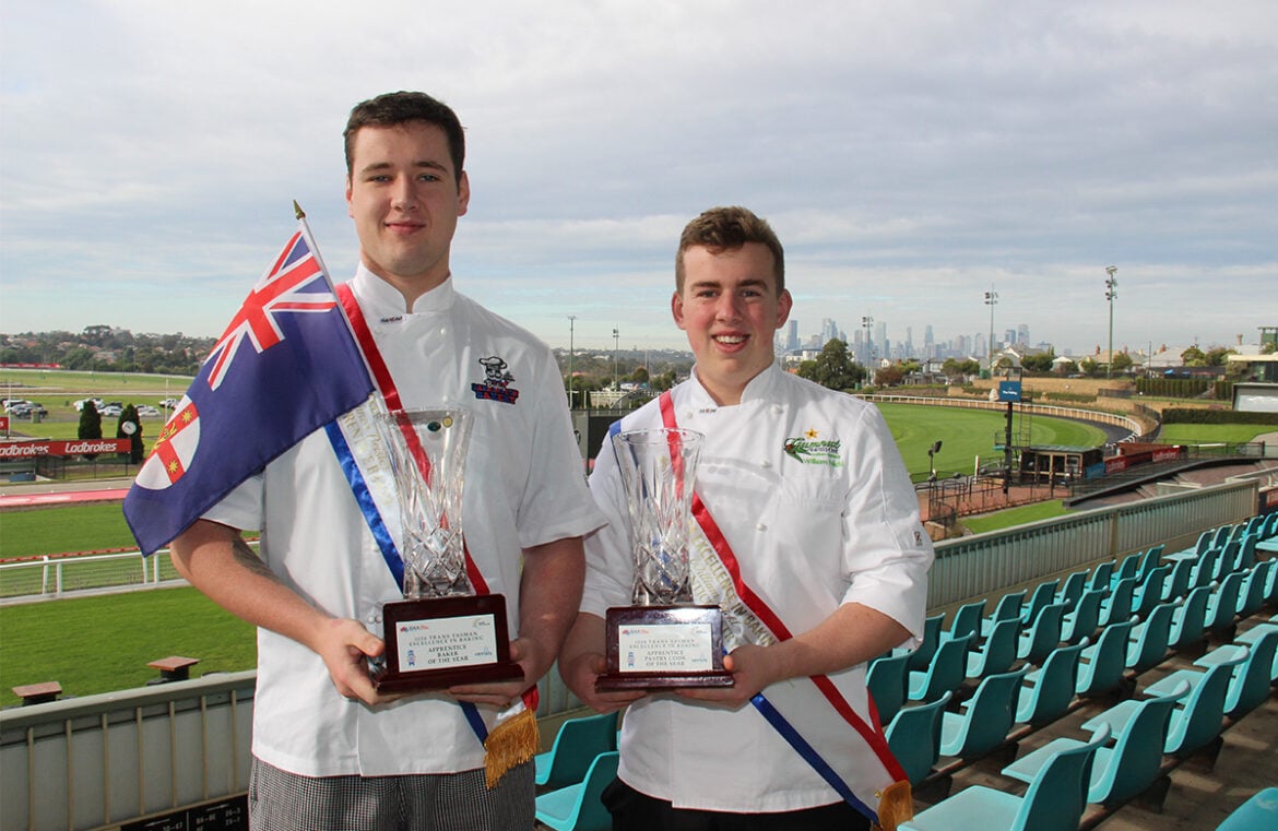 Connor Beisler and William Nickl stand in the grandstands of a horse racecourse. They have green plastic seats around them and the grass track in the background. Both men wear white chef shirts and are holding wooden trophies. They are smiling at the camera.