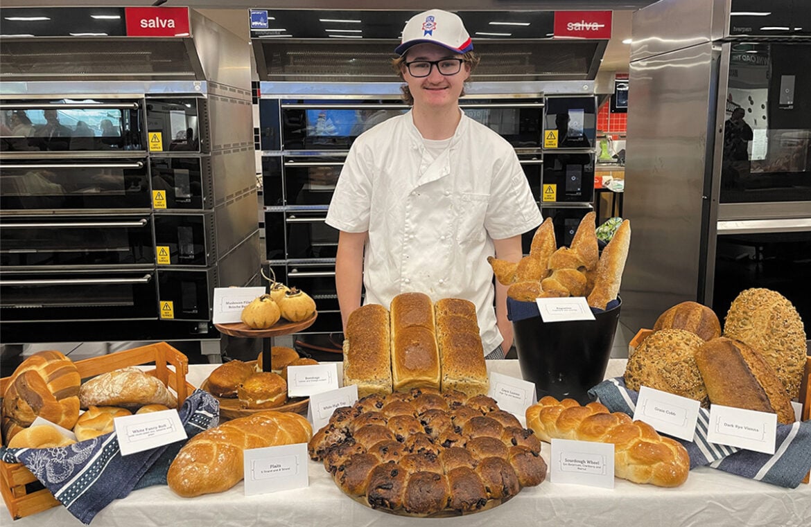 ABINSA winner Caleb Brazsell poses in a white apron behind a table covered with different types of bread. Caleb has short brown hair and is wearing a cap and glasses.