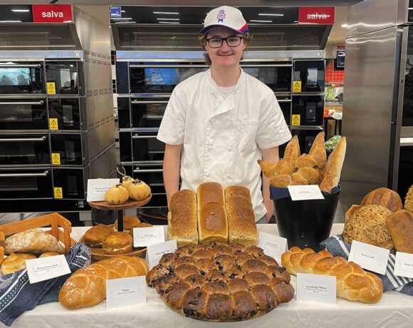 ABINSA winner Caleb Brazsell poses in a white apron behind a table covered with different types of bread. Caleb has short brown hair and is wearing a cap and glasses.