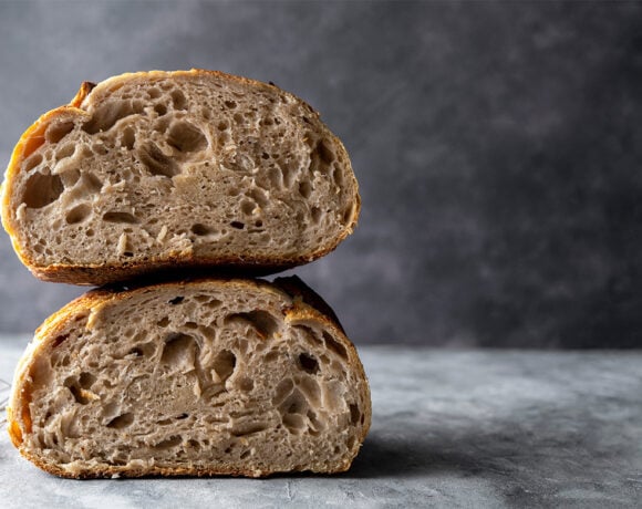 A loaf of sourdough bread is cut in half, with the halves stacked on top of each other to show the interior. The bread sits on a grey table with a dark grey wall behind.