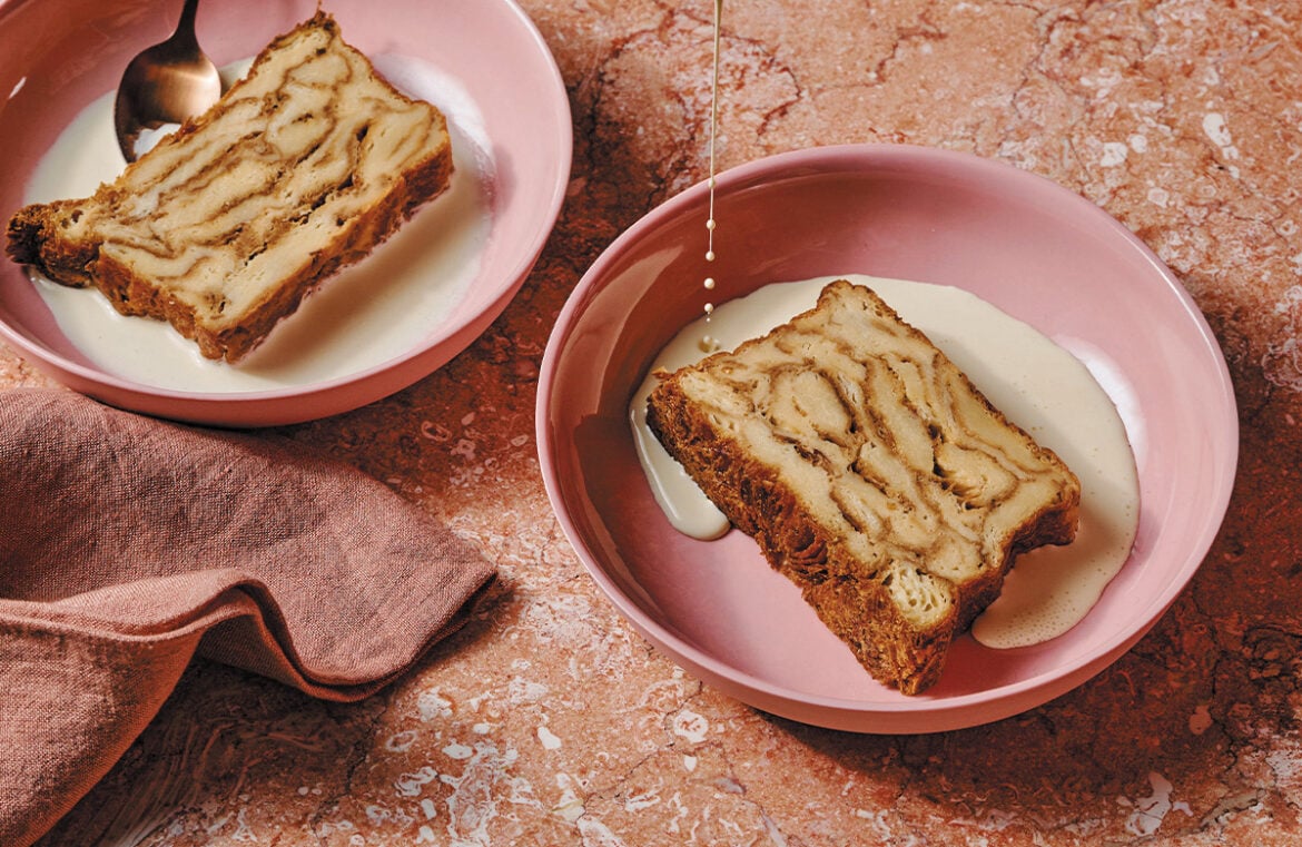 Two pink bowls sit on a marbled pink counter. In each bowl is a slice of bread and butter pudding. A hand reaches in and pours custard over one of the bowls.