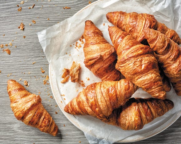 Croissants sit on baking paper. One sits by itself to the left on a grey table