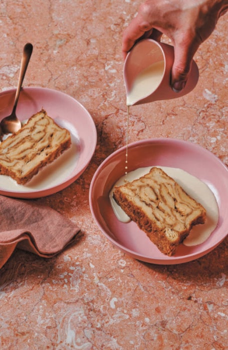 Croissant bread and butter pudding by Lune. Pictured are two pink bowls. In each is a slice of bread and butter pudding. A hand reaches on from the top right with a cream jug holding custard that is being poured over the top.