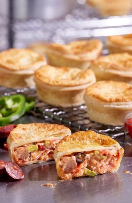 EOI beef and pepperoni pies sit on a stainless steel bench. One is cut in half to show the filling.