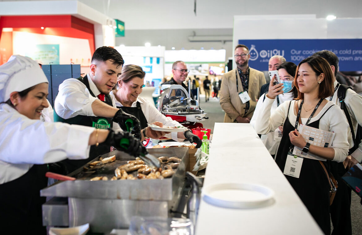 Fine Food Australia 2024. Pictured are attendees looking over a counter and watching on as three chefs work on a hot stove.