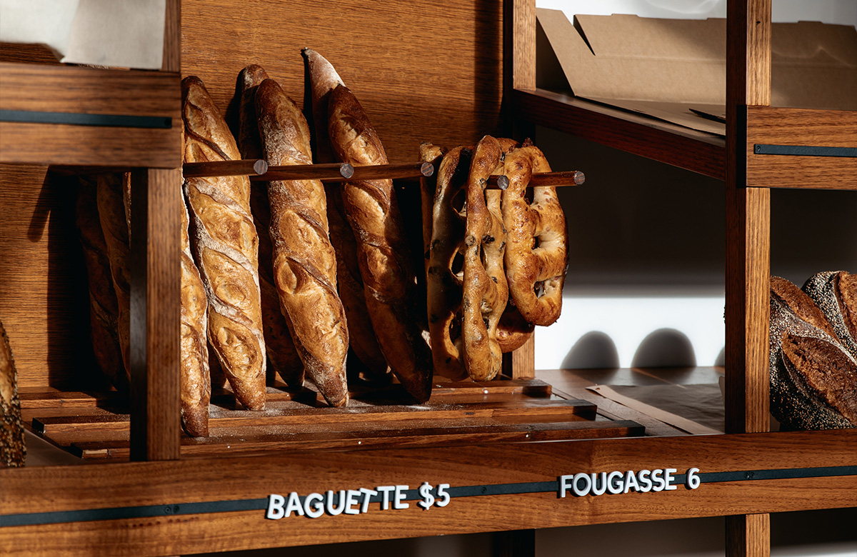 Pictured is baguettes and fougasse. They are on a wooden shelf with white labels underneath.