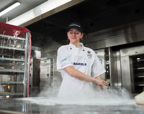 Pictured is Imogen Fearon. She is wearing chef whites and a dark cap. Her hair is tied back and she has a smile on her face. She's looking down as she sprinkles flour onto a stainless steel bench.