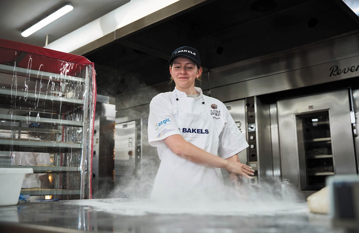 Pictured is Imogen Fearon. She is wearing chef whites and a dark cap. Her hair is tied back and she has a smile on her face. She's looking down as she sprinkles flour onto a stainless steel bench.