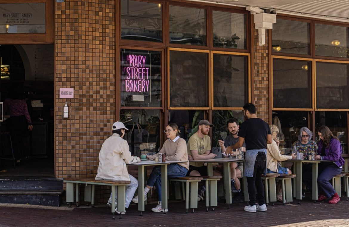 Pictured is the exterior of Mary Street Bakery. The store has dark bricks and ground to ceiling windows along one side. Patrons are sitting on tables on the sidewalk and a neon pink sign in a window says Mary Street Bakery