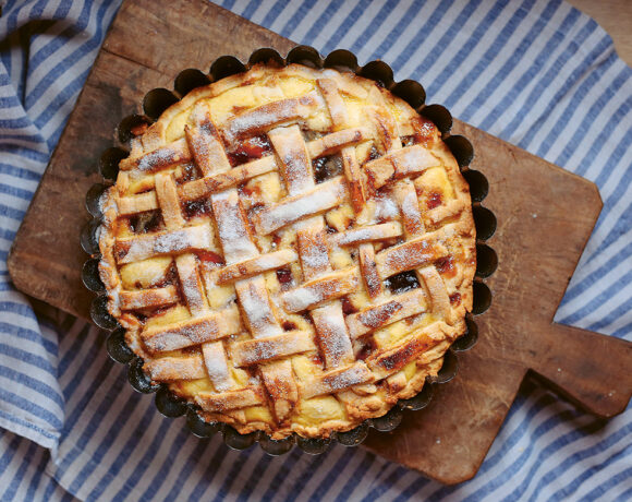 Plum and ricotta crostata is pictured on blue and white striped cloth. It sits on a wooden paddle cuttle board.