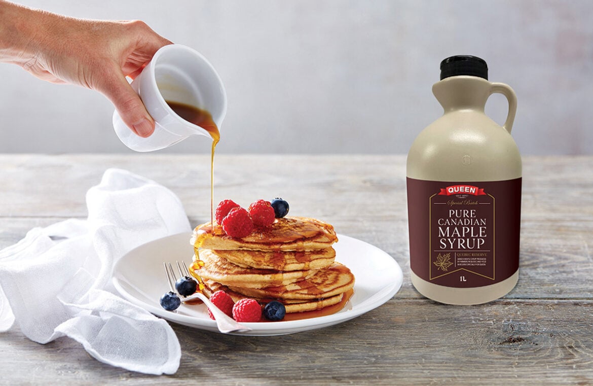 A bottle of Queen Professional maple syrup sits to the right of a plate pancakes. The pancakes have strawberries and blueberries scattered on top, and a hand reaches in, holding a white jug of maple syrup that is being poured over the pancakes