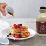 A bottle of Queen Professional maple syrup sits to the right of a plate pancakes. The pancakes have strawberries and blueberries scattered on top, and a hand reaches in, holding a white jug of maple syrup that is being poured over the pancakes