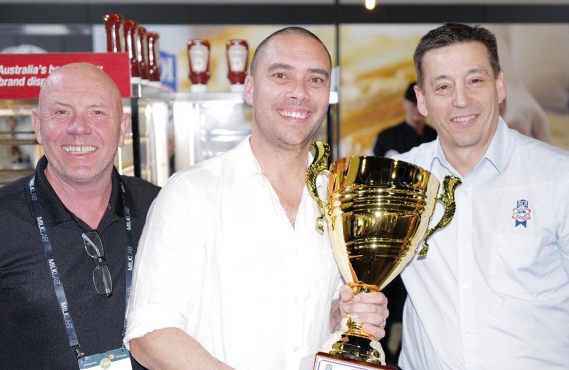 Pictured is Nathan Williams from Rolling Pin Pies and Cakes. He was a major winner at the 2023 The Official Great Aussie Pie competition. Nathan is a bald man, he's holding a large gold trophy and is wearing a white polo shirt. He's smiling at the camera.