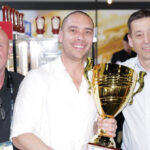 Pictured is Nathan Williams from Rolling Pin Pies and Cakes. He was a major winner at the 2023 The Official Great Aussie Pie competition. Nathan is a bald man, he's holding a large gold trophy and is wearing a white polo shirt. He's smiling at the camera.