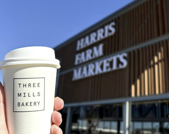 Three Mills Bakery has opened its newest store in Majura Park. Pictured is a white disposable coffee cup with a black Three Mills Bakery logo on it. Out of focus in the background is the exterior of the Harris Farm Markets store and signage.