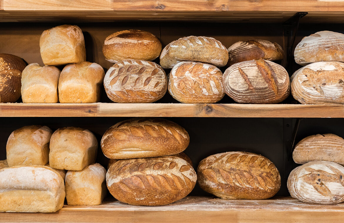 2024 baking trends. Pictured is a wooden shelf with two levels. Each levels is full of freshly baked bread loaves. The loaves are all golden brown and unpackaged. There is a light dusting of flour on the shelves under the bread.