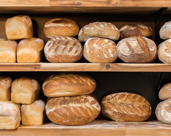 2024 baking trends. Pictured is a wooden shelf with two levels. Each levels is full of freshly baked bread loaves. The loaves are all golden brown and unpackaged. There is a light dusting of flour on the shelves under the bread.