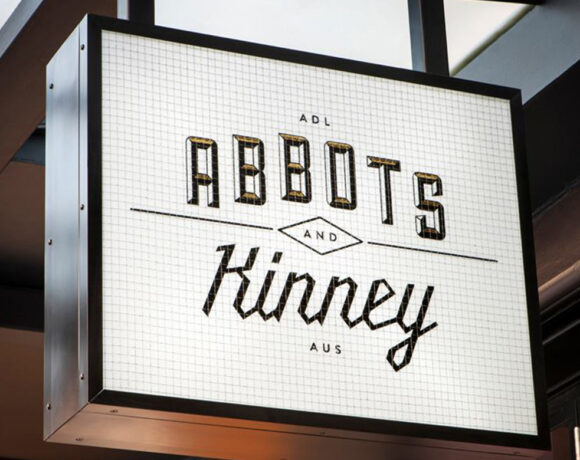 Abbots and Kinney sign. It's a white rectangle with Abbots and Kinney written in black writing on it. It's suspended from the ceiling by two metal poles.