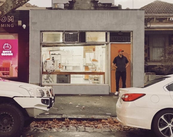 A man in black clothing and a rec cap stands at the door to Amann Patisserie. The building is grey concrete and has a large window across the front. You can see the kitchen through the window.