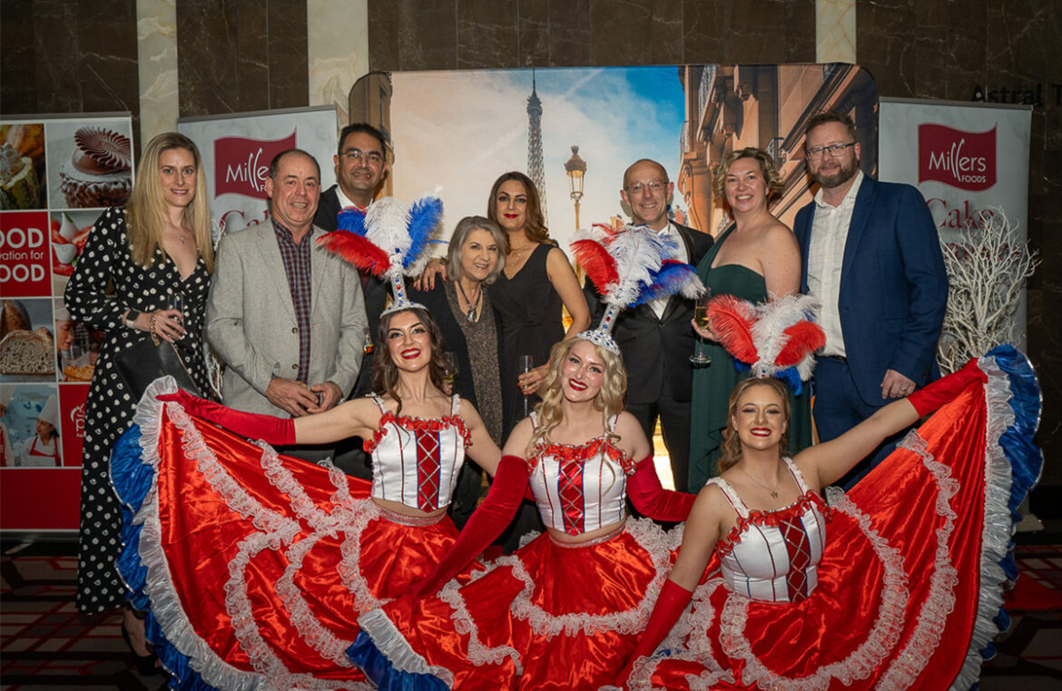 A group of people stand in front of a poster of the Eiffel Tower. In front of them are three women dressed in blue, red and white costumes from teh Moulin Rouge. They're celebrating the BIEWA gala awards