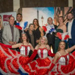 A group of people stand in front of a poster of the Eiffel Tower. In front of them are three women dressed in blue, red and white costumes from teh Moulin Rouge. They're celebrating the BIEWA gala awards