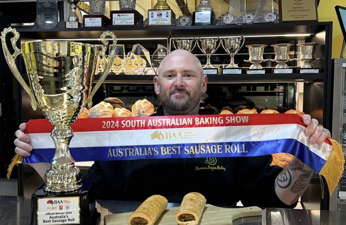 Pictured is Jason Spencer from Banana Boogie Bakery. Jason is a while man with a bald head and facial hair. He stands behind a counter, holding a ribbon that says Australia's Best Sausage Roll. Next to him is a gold trophy that also says Australia's Best Sausage Roll. He has two sausage rolls sitting on the counter in front of him.