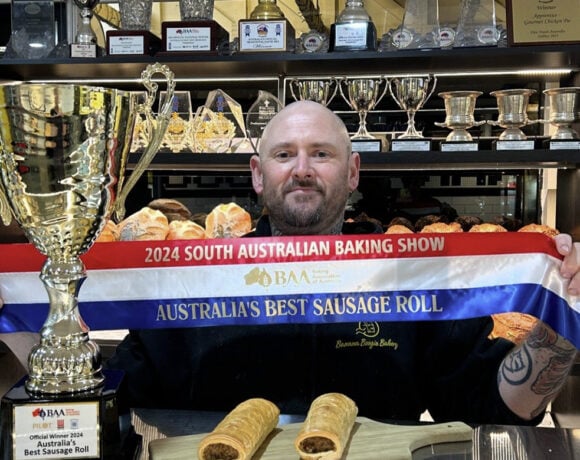 Pictured is Jason Spencer from Banana Boogie Bakery. Jason is a while man with a bald head and facial hair. He stands behind a counter, holding a ribbon that says Australia's Best Sausage Roll. Next to him is a gold trophy that also says Australia's Best Sausage Roll. He has two sausage rolls sitting on the counter in front of him.
