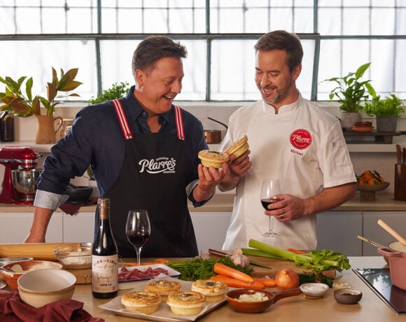 Darren Purchese is pictured with Ferguson Plarre CEO Steve Plarre at a kitchen bench. The bench is covered with fresh produce, and has an open bottle of wine alongside 2 wine glasses on it.