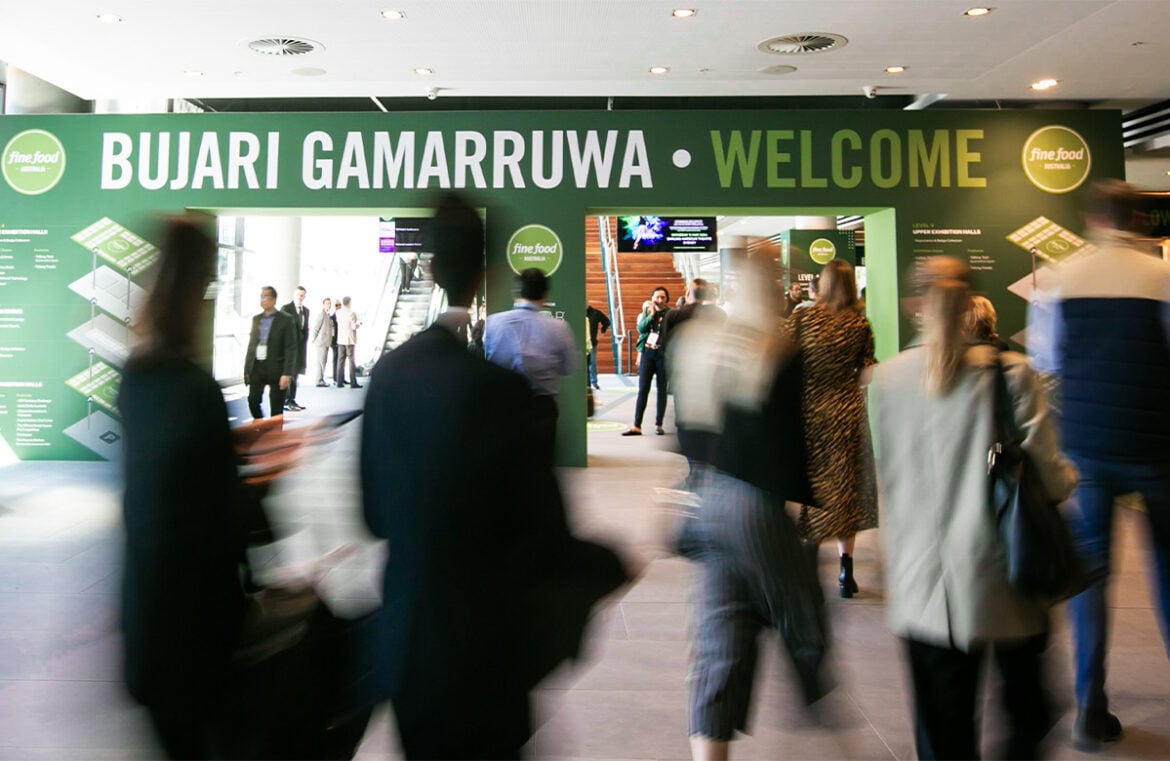 A blurred entryway to Fine Food Australia 2024. The green sign is running across teh top of the image with people blurred while walking past it.