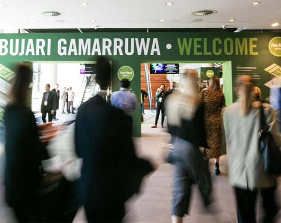 A blurred entryway to Fine Food Australia 2024. The green sign is running across teh top of the image with people blurred while walking past it.