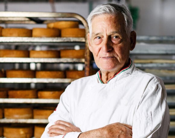 Giorgio Angele an older man with white hair neatly brushed back, stands in a white chef's shirt. His arms are crossed across his chest and he's looking at the camera with a serious expression. Behind him is a tall tower filled with baked goods.