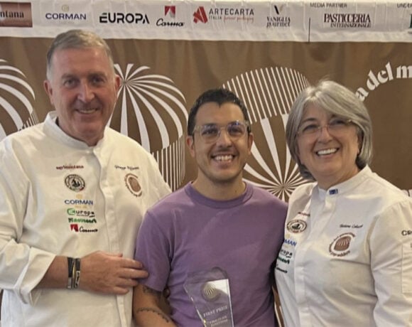 Jonny Pisanelli from Sonoma took out the chocolate panettone category. Picutred is Jonny. He woears a purple t-shirt and hold a glass trophy in front of him. He as short dark hair and is smiling at the camera. Next to him are two bakers in chefs whites.