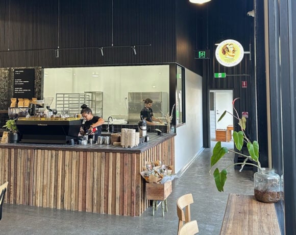 The Bread Social's Currumbin Waters bakery cafe. Picutred is a wooden counter with a floor to ceiling galss wall behind it. A woman stands at the counter, wearing a black shirt and making coffee. Behind her a baker stands a bench making bread. He also wears a black shirt.