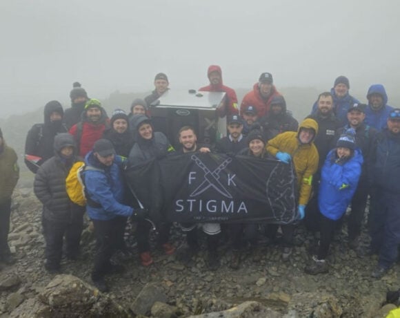 Pictured is the 25 team member from Unox UK. They all wear wet weather hiking gear and stand in a group on a rock. They are surrounded by dark grey clouds. The team are holding a black sign with white writing that says F**k Stigma on it. The missing letters in the word have been replaced by can image of crossed kitchen knives.