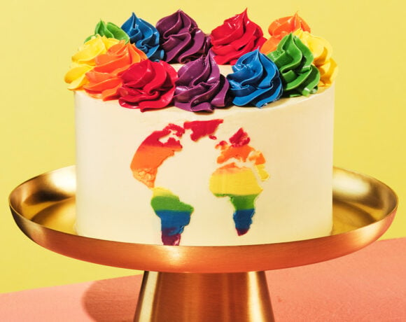 World Pride cake sits on a golden cake stand with a yellow wall behind it. The cake is voered with vanilla icing and has rainbow coloured swirls on teh top.