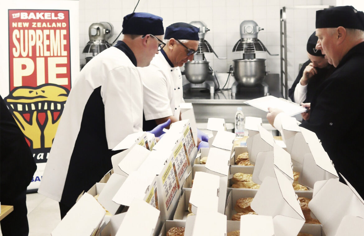 Four men gather around a table filled with white boxes. The two men on the left are in chef whites with black hats, the two men on the right are in black shirts. They lean over the table towards each other, looking at the pies in between them. They're the judges for the Bakels NZ Supreme Pie Award.
