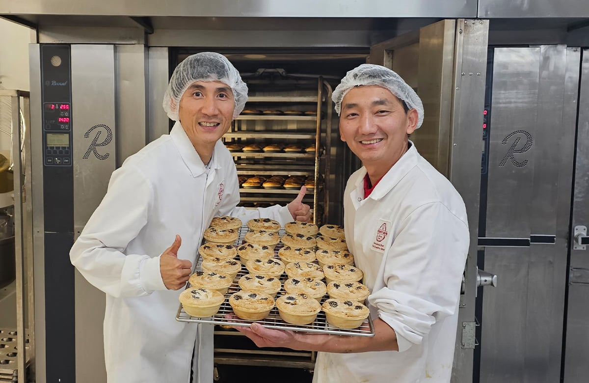 Teo men stand in a kitchen. One hold a tray of meat pies, the other gives the thumbs up to the camera. 