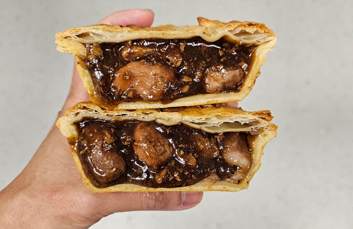 A meat pie is cut in half, with the halves stacked on top of each other. A hand holds them in front of a white wall