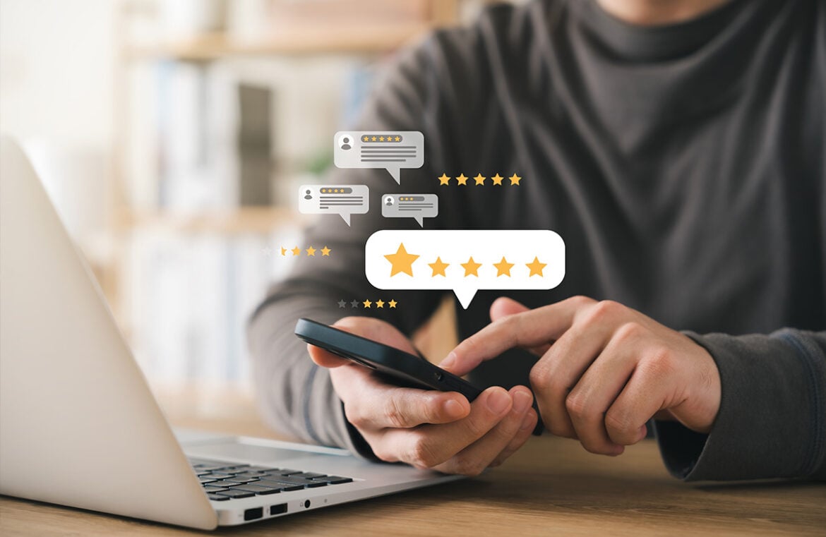 A man sits at a desk. He has an open laptop in front of him and a phone in his hand. HE has stars around his hand, signalling he's leaving a customer review